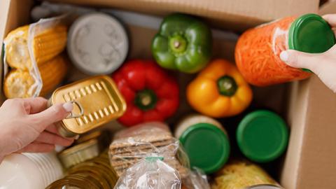 A person filling a cardboard box with food items items such as bell peppers, corn on the cob, sardines and crackers among various other items.