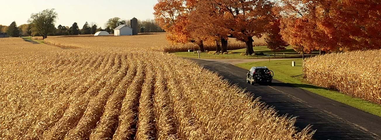 A single car driving down a country road past a field of crops and a farm.