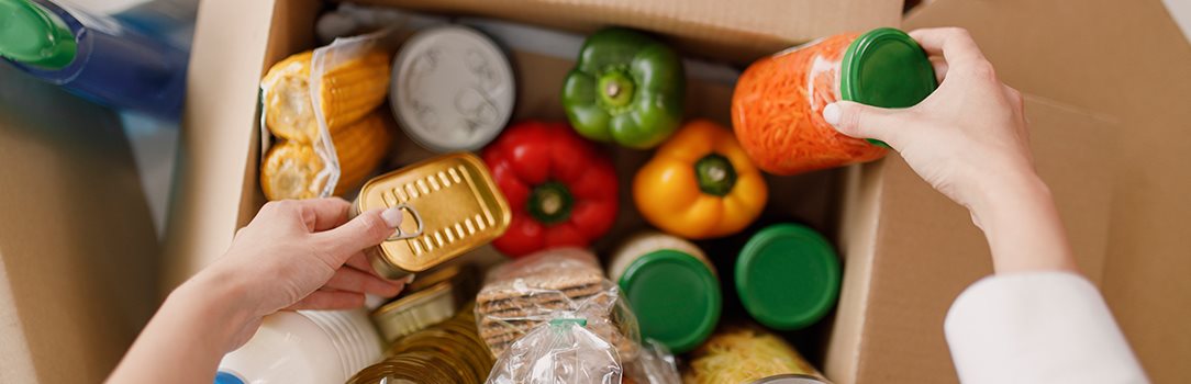 A person filling a cardboard box with food items items such as bell peppers, corn on the cob, sardines and crackers among various other items.