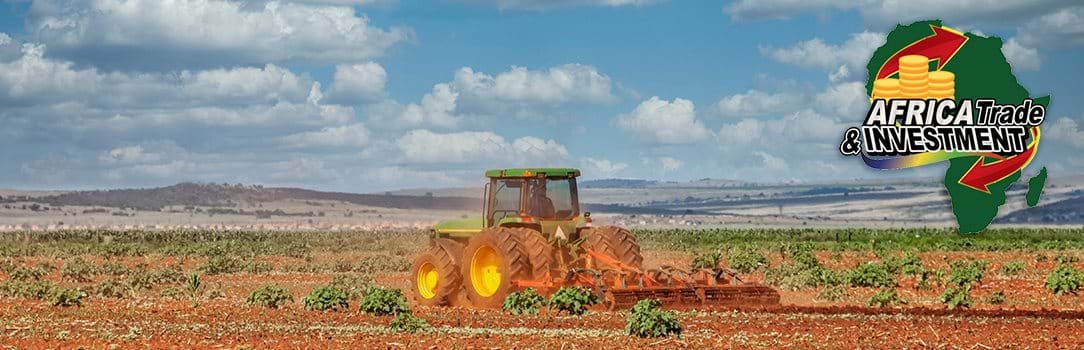 Photo of a green tractor plowing an agricultural field with low mountains and a cloudy, blue sky in the background.
