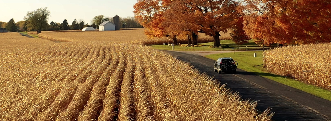 A single car driving down a country road past a field of crops and a farm.